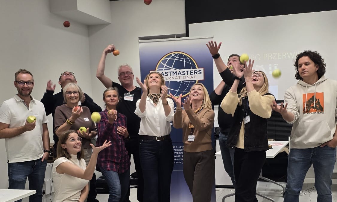 Uczestnicy spotkania Toastmasters Opole podrzucają jabłka przed banerem Toastmasters International