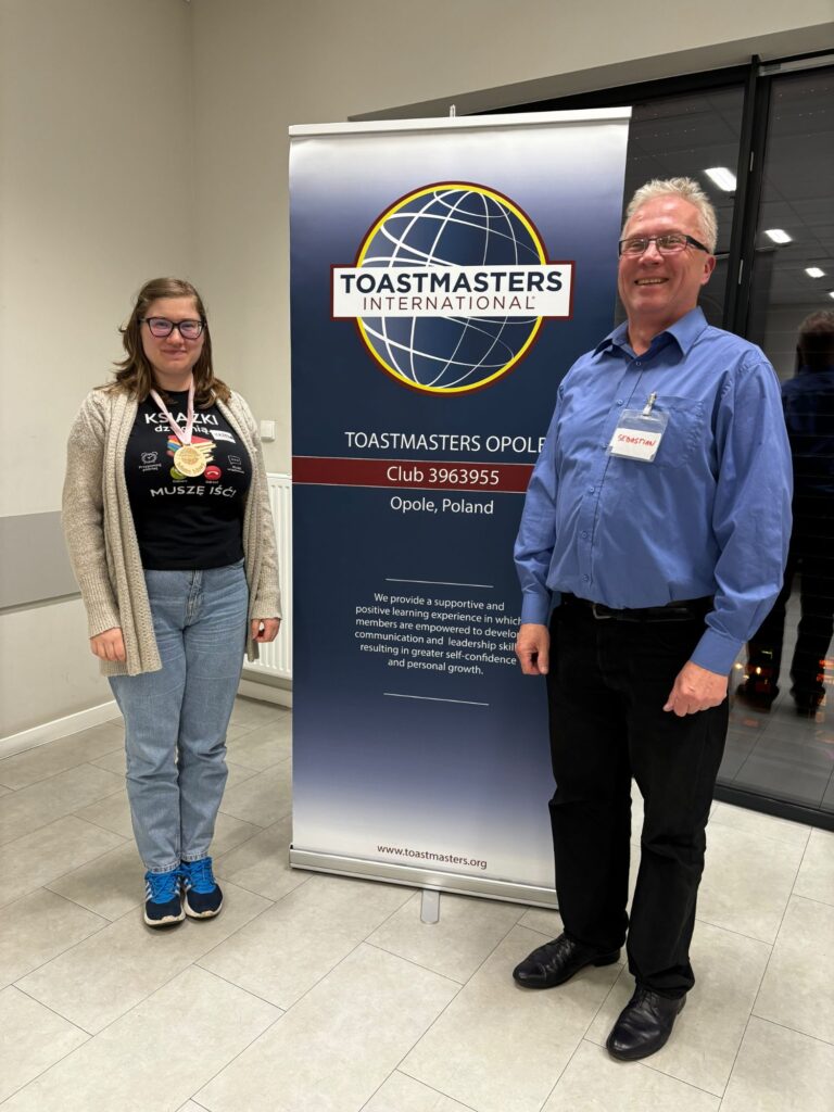 Martyna z medalem za najlepszą improwizację i Sebastian, moderator gorących pytań, przed banerem Toastmasters Opole
